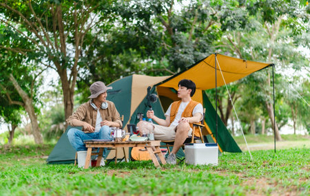 Two Asian male friends enjoy relaxing moment together at campsite surrounded by nature. With a tent set up and gear around them, the atmosphere reflects leisure, companionship, and outdoor freedomの写真素材