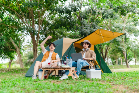 Two Asian male friends enjoy relaxing moment together at campsite surrounded by nature. With a tent set up and gear around them, the atmosphere reflects leisure, companionship, and outdoor freedomの写真素材