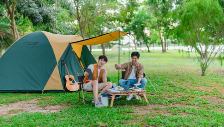 Two Asian male friends enjoy relaxing moment together at campsite surrounded by nature. With a tent set up and gear around them, the atmosphere reflects leisure, companionship, and outdoor freedomの写真素材