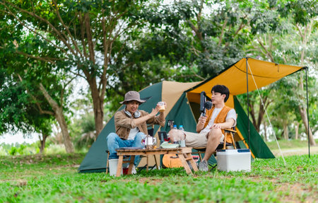 Two Asian male friends enjoy relaxing moment together at campsite surrounded by nature. With a tent set up and gear around them, the atmosphere reflects leisure, companionship, and outdoor freedomの写真素材