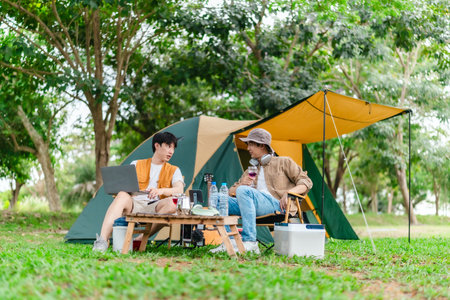 Two Asian male friends enjoy relaxing moment together at campsite surrounded by nature. With a tent set up and gear around them, the atmosphere reflects leisure, companionship, and outdoor freedomの写真素材