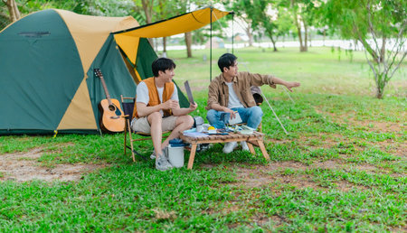 Two Asian male friends enjoy relaxing moment together at campsite surrounded by nature. With a tent set up and gear around them, the atmosphere reflects leisure, companionship, and outdoor freedomの写真素材