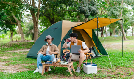 Two Asian male friends enjoy relaxing moment together at campsite surrounded by nature. With a tent set up and gear around them, the atmosphere reflects leisure, companionship, and outdoor freedomの写真素材