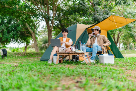 Two Asian male friends enjoy relaxing moment together at campsite surrounded by nature. With a tent set up and gear around them, the atmosphere reflects leisure, companionship, and outdoor freedomの写真素材