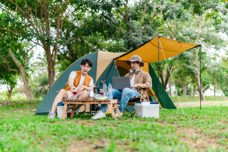 Two Asian male friends enjoy relaxing moment together at campsite surrounded by nature. With a tent set up and gear around them, the atmosphere reflects leisure, companionship, and outdoor freedomの写真素材