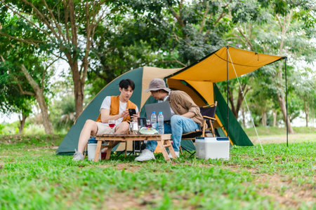 Two Asian male friends enjoy relaxing moment together at campsite surrounded by nature. With a tent set up and gear around them, the atmosphere reflects leisure, companionship, and outdoor freedomの写真素材