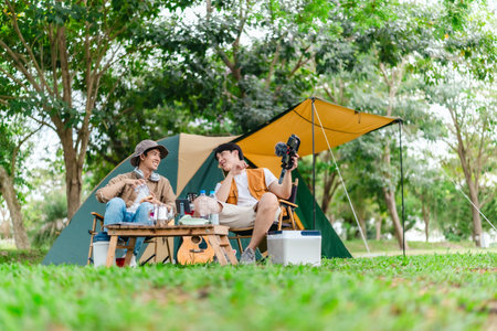 Two Asian male friends enjoy relaxing moment together at campsite surrounded by nature. With a tent set up and gear around them, the atmosphere reflects leisure, companionship, and outdoor freedomの写真素材