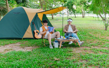 Two Asian male friends enjoy relaxing moment together at campsite surrounded by nature. With a tent set up and gear around them, the atmosphere reflects leisure, companionship, and outdoor freedomの写真素材