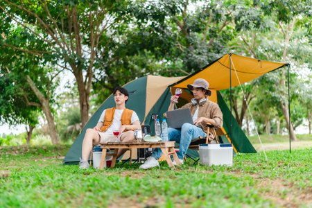 Two Asian male friends enjoy relaxing moment together at campsite surrounded by nature. With a tent set up and gear around them, the atmosphere reflects leisure, companionship, and outdoor freedomの写真素材