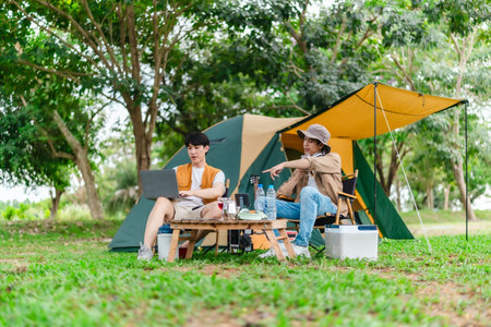 Two Asian male friends enjoy relaxing moment together at campsite surrounded by nature. With a tent set up and gear around them, the atmosphere reflects leisure, companionship, and outdoor freedomの写真素材