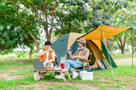 Two Asian male friends enjoy relaxing moment together at campsite surrounded by nature. With a tent set up and gear around them, the atmosphere reflects leisure, companionship, and outdoor freedomの写真素材