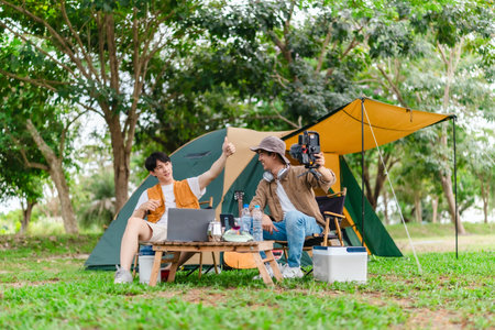 Two Asian male friends enjoy relaxing moment together at campsite surrounded by nature. With a tent set up and gear around them, the atmosphere reflects leisure, companionship, and outdoor freedomの写真素材