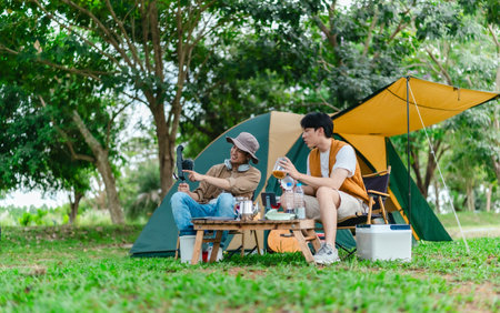 Two Asian male friends enjoy relaxing moment together at campsite surrounded by nature. With a tent set up and gear around them, the atmosphere reflects leisure, companionship, and outdoor freedomの写真素材