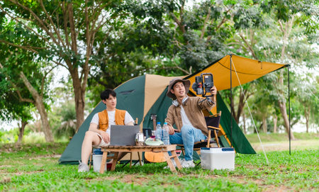 Two Asian male friends enjoy relaxing moment together at campsite surrounded by nature. With a tent set up and gear around them, the atmosphere reflects leisure, companionship, and outdoor freedomの写真素材