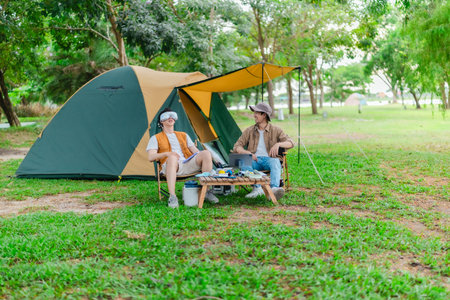Two Asian male friends enjoy relaxing moment together at campsite surrounded by nature. With a tent set up and gear around them, the atmosphere reflects leisure, companionship, and outdoor freedomの写真素材