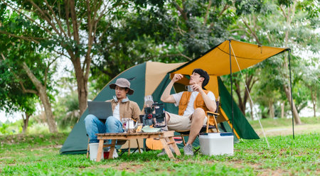 Two Asian male friends enjoy relaxing moment together at campsite surrounded by nature. With a tent set up and gear around them, the atmosphere reflects leisure, companionship, and outdoor freedomの写真素材
