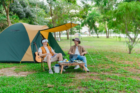 Two Asian male friends enjoy relaxing moment together at campsite surrounded by nature. With a tent set up and gear around them, the atmosphere reflects leisure, companionship, and outdoor freedomの写真素材