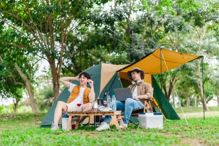 Two Asian male friends enjoy relaxing moment together at campsite surrounded by nature. With a tent set up and gear around them, the atmosphere reflects leisure, companionship, and outdoor freedomの写真素材