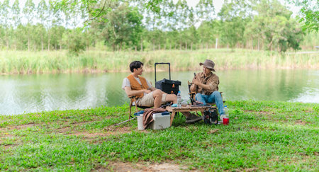 Two Asian male friends enjoy relaxing moment together at campsite surrounded by nature. With a tent set up and gear around them, the atmosphere reflects leisure, companionship, and outdoor freedomの写真素材