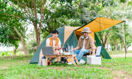 Two Asian male friends enjoy relaxing moment together at campsite surrounded by nature. With a tent set up and gear around them, the atmosphere reflects leisure, companionship, and outdoor freedomの写真素材