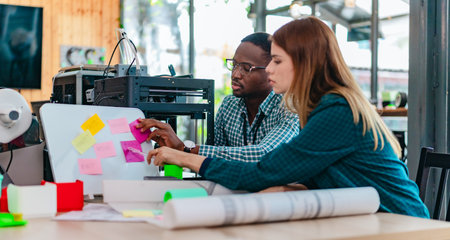 A diverse team of startup professionals collaborates on a 3D printing project in a modern workspace....