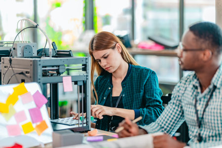 A diverse team of startup professionals collaborates on a 3D printing project in a modern workspace....