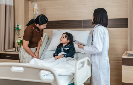 Caring Asian female doctor checks on young girl in hospital bed during consultation, while child's mother provide support and comfort. warmth, trust, and professional medical care in clinical settingの写真素材