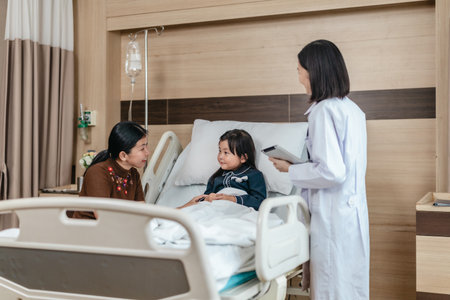 Caring Asian female doctor checks on young girl in hospital bed during consultation, while child's...