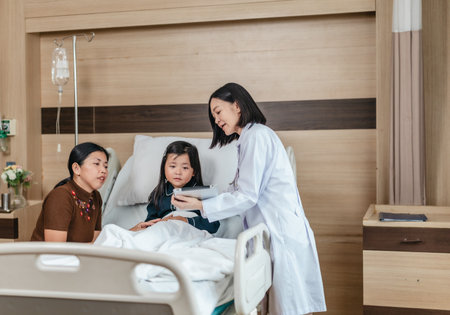 Caring Asian female doctor checks on young girl in hospital bed during consultation, while child's mother provide support and comfort. warmth, trust, and professional medical care in clinical settingの写真素材