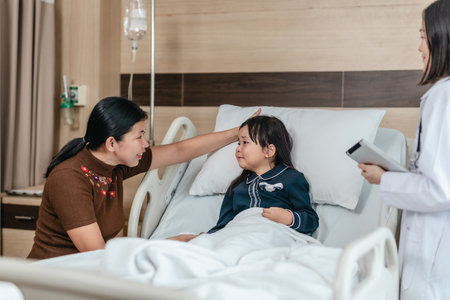 Caring Asian female doctor checks on young girl in hospital bed during consultation, while child's mother provide support and comfort. warmth, trust, and professional medical care in clinical settingの写真素材