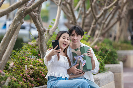 An Asian couple shares a joyful moment while exchanging gift boxes in a scenic outdoor setting. The...