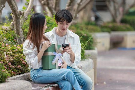 An Asian couple shares a joyful moment while exchanging gift boxes in a scenic outdoor setting. The scene captures feelings of love, celebration, and connection during a special occasion.の写真素材