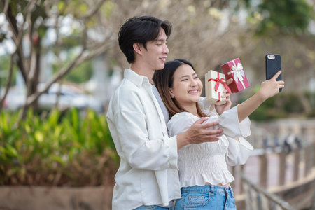 An Asian couple shares a joyful moment while exchanging gift boxes in a scenic outdoor setting. The scene captures feelings of love, celebration, and connection during a special occasion.の写真素材