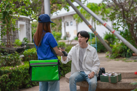 Asian man uses smartphone to make cashless payment while receiving delivery from female courier....