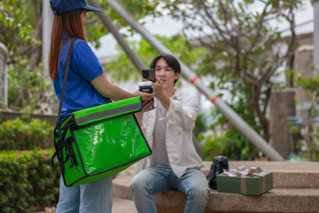 Asian man uses smartphone to make cashless payment while receiving delivery from female courier. Modern transaction takes place outdoors, capturing convenience and efficiency of contactless service.の写真素材