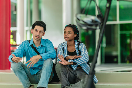 Two university students from different cultural backgrounds sit on campus step, engaged in friendly...