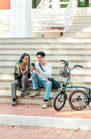 Two diverse college friends sit on sunlit campus steps, consulting a smartphone beside a compact folding bicycle and helmet, illustrating eco friendly urban commuting and modern student life.の写真素材