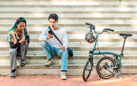 Two diverse college friends sit on sunlit campus steps, consulting a smartphone beside a compact...