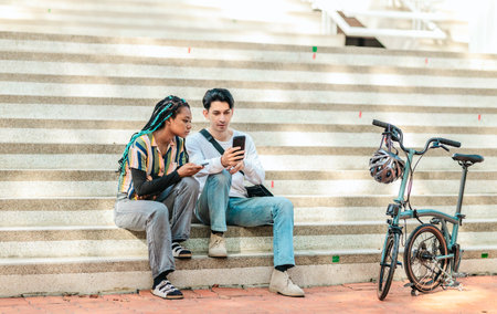 Two diverse college friends sit on sunlit campus steps, consulting a smartphone beside a compact...