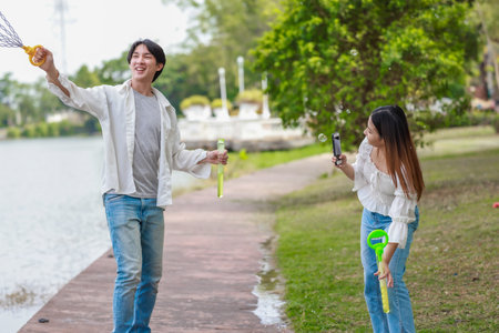 A cheerful Asian couple enjoys blowing bubbles by a lakeside path on a sunny day. The joyful moment...