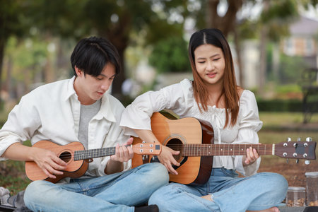 Asian couple enjoys relaxing day at park, smiling and playing guitar and ukulele together during a picnic. Capturing a joyful and candid moment of connection, music, and leisure in natural setting.の写真素材