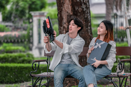 Asian couple enjoys heartfelt moment on park bench as man presents bouquet of red flowers and gift...