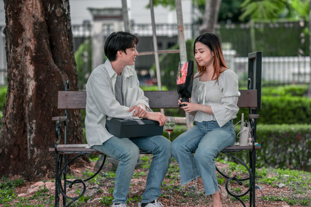 Asian couple enjoys heartfelt moment on park bench as man presents bouquet of red flowers and gift box to his partner. The warm, outdoor setting reflects love, appreciation, and romantic celebrationの写真素材