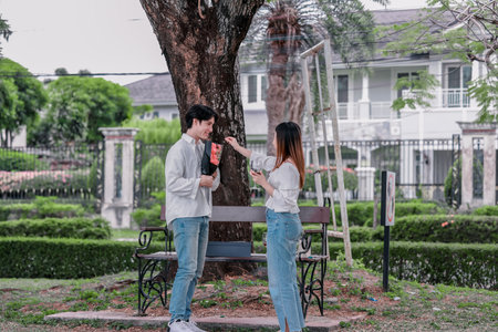 Asian couple enjoys heartfelt moment on park bench as man presents bouquet of red flowers and gift box to his partner. The warm, outdoor setting reflects love, appreciation, and romantic celebrationの写真素材