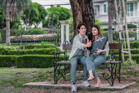 Asian couple enjoys heartfelt moment on park bench as man presents bouquet of red flowers and gift box to his partner. The warm, outdoor setting reflects love, appreciation, and romantic celebrationの写真素材