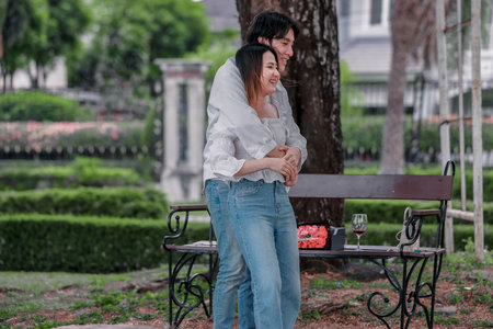 Asian couple enjoys heartfelt moment on park bench as man presents bouquet of red flowers and gift box to his partner. The warm, outdoor setting reflects love, appreciation, and romantic celebrationの写真素材