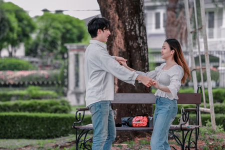 Asian couple enjoys heartfelt moment on park bench as man presents bouquet of red flowers and gift box to his partner. The warm, outdoor setting reflects love, appreciation, and romantic celebrationの写真素材