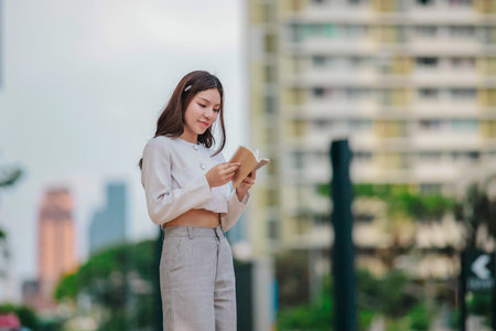 Asian woman dressed in modern business casual attire is seen writing in notebook while standing outdoors in urban setting. Conveying sense of focus, productivity, and creativity in city environmentの写真素材