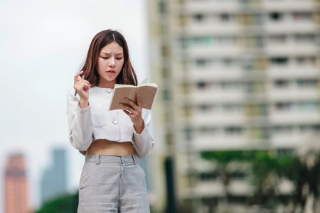 Asian woman dressed in modern business casual attire is seen writing in notebook while standing outdoors in urban setting. Conveying sense of focus, productivity, and creativity in city environmentの写真素材