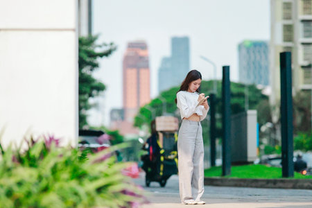 Asian woman dressed in modern business casual attire is seen writing in notebook while standing outdoors in urban setting. Conveying sense of focus, productivity, and creativity in city environmentの写真素材
